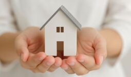 Modelo de casa blanca con tejado gris sostenido cuidadosamente por unas manos humanas, simbolizando protección y propiedad inmobiliaria